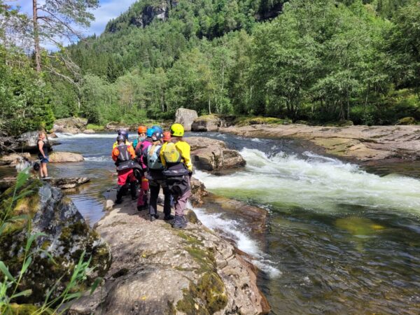 En gruppe står langs elvebredden og ser på elven. De retter oppmerksomheten mot vannet og landskapet rundt, mens de diskuterer og peker på ulike trekk ved elven.