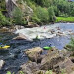 Deltakerne på kurset navigerer gjennom stryk og raskt flytende vann mens de padler nedover elven. De viser god kontroll over kajakkene sine og samarbeider for å manøvrere trygt gjennom utfordrende vannforhold.