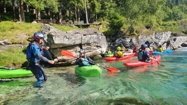 En gruppe deltakere på et grunnkurs i elvepaddling er klare til å sette ut i elven. De har på seg redningsvester og har kajakkene klare ved vannkanten.